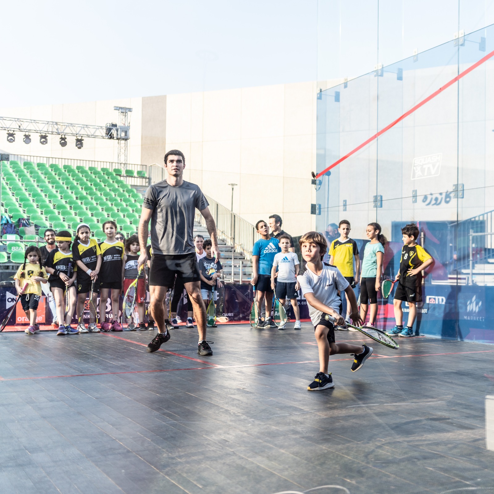 A group of children stood behind a professional male squash player and a young boy playing squash in an outdoor squash court